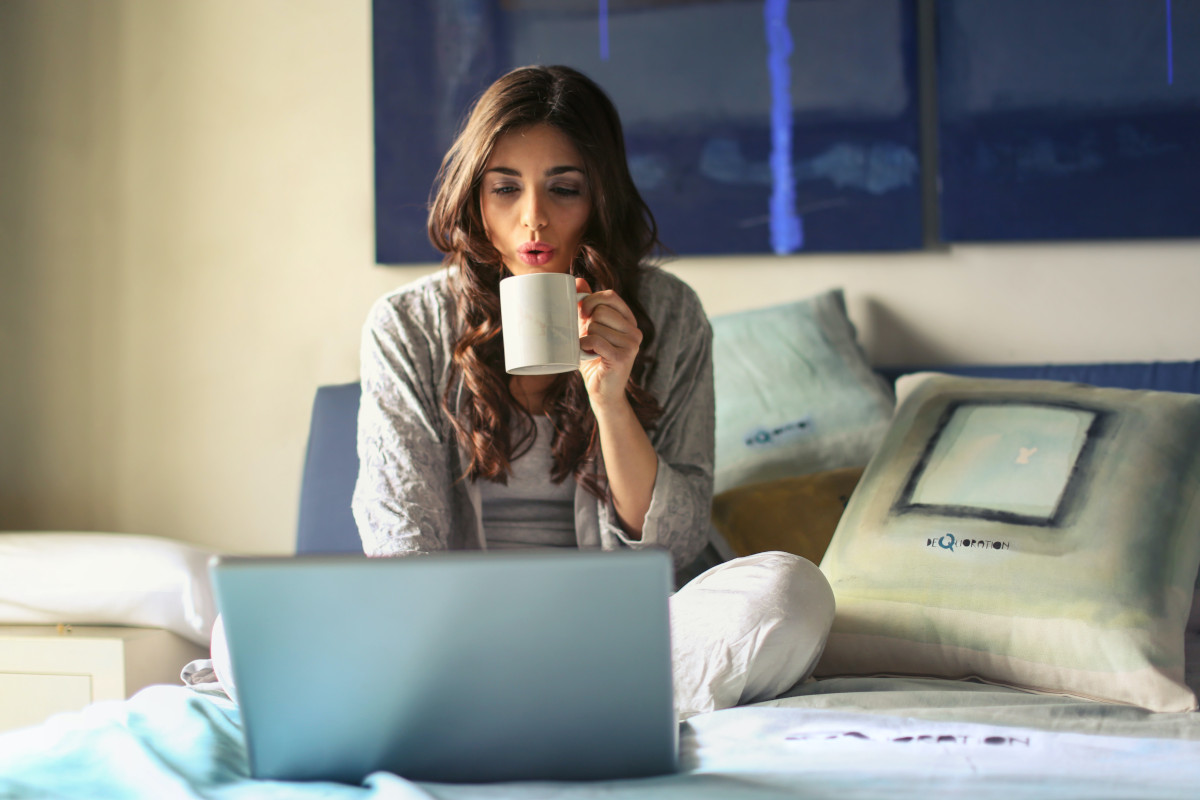 A women working remotely from her laptop while sitting in bed and blowing on a hot cup of coffee.